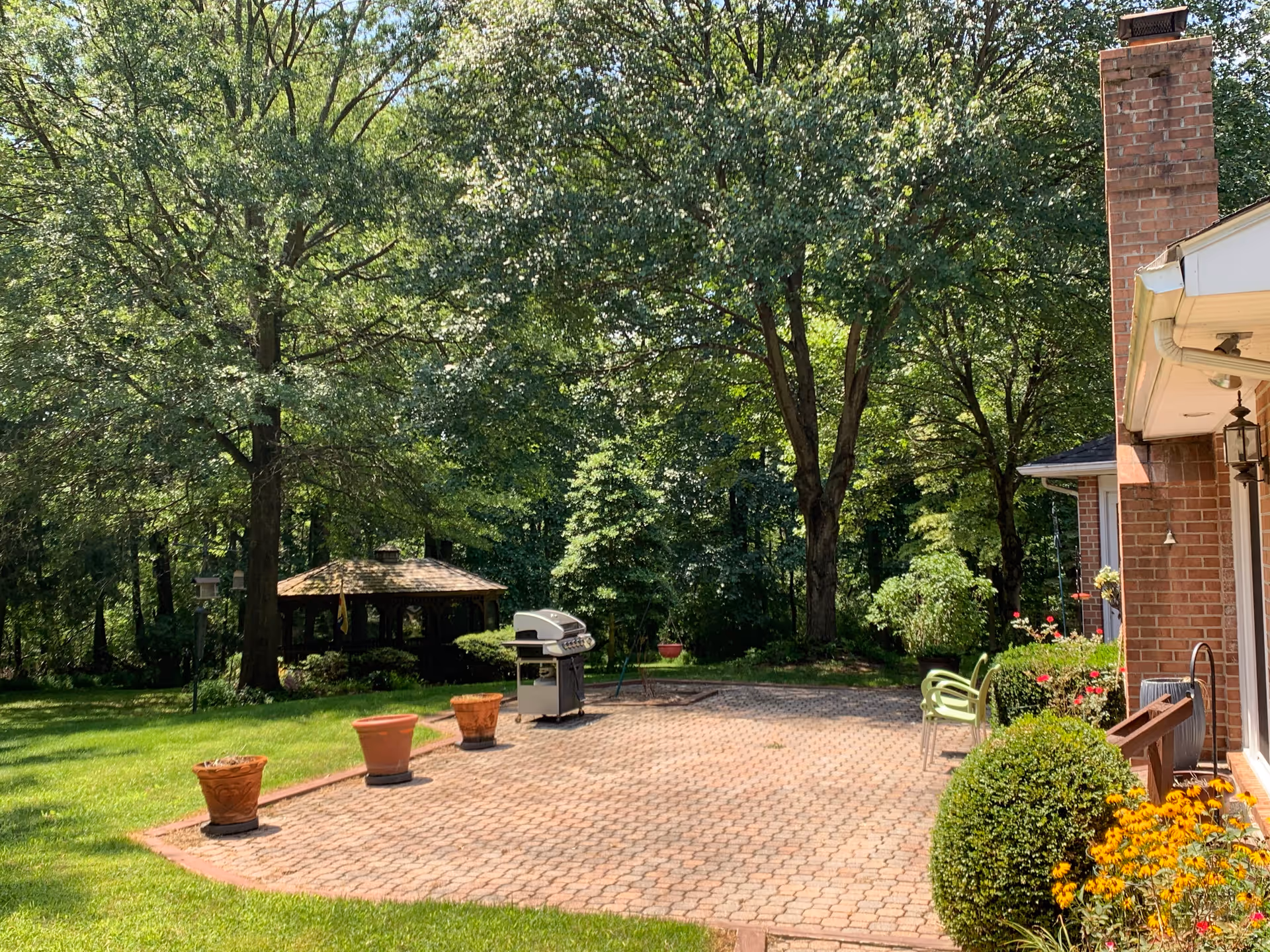 A sunny outdoor patio area with a brick paved floor, several empty flower pots, a barbecue grill, and green chairs. The patio is adjacent to a brick building with a chimney. Surrounding the patio are green bushes, yellow flowers, and large trees providing shade. In the background, there is a wooden gazebo nestled among the trees.