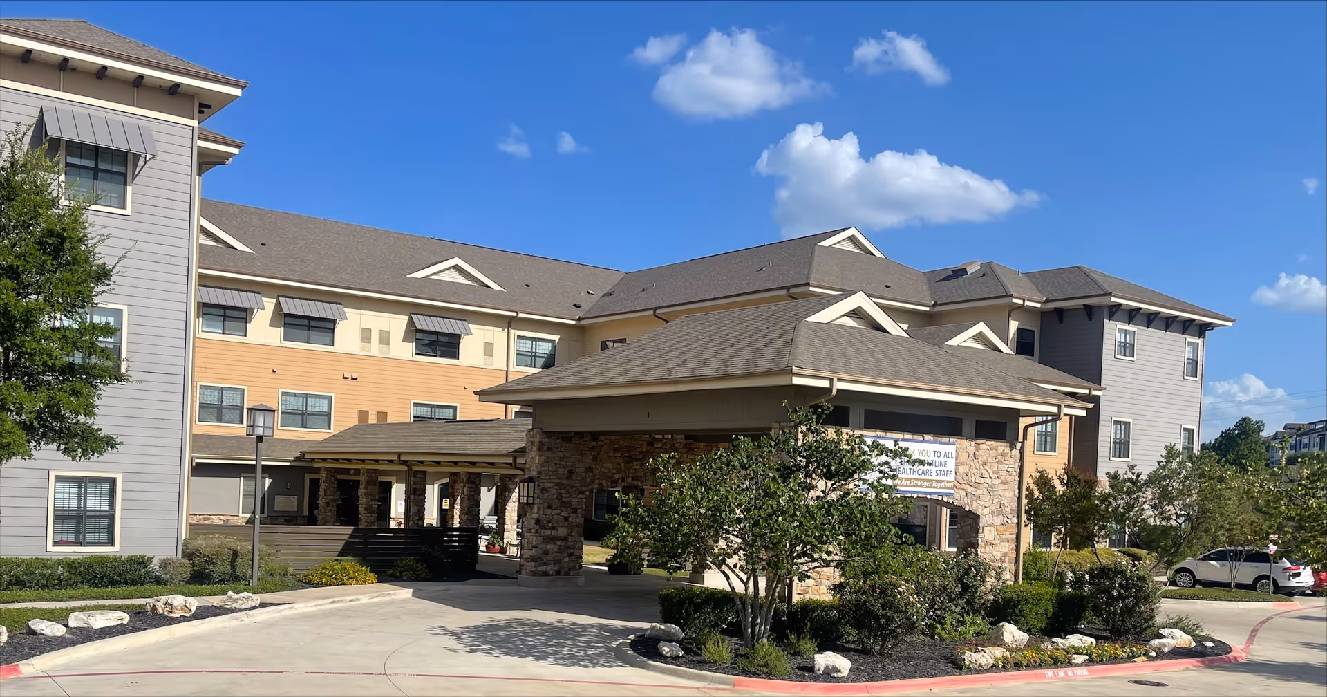 Exterior view of a multi-story senior living facility building with beige and gray siding, a covered entrance supported by stone pillars, landscaped bushes and trees, and a clear blue sky with some clouds.