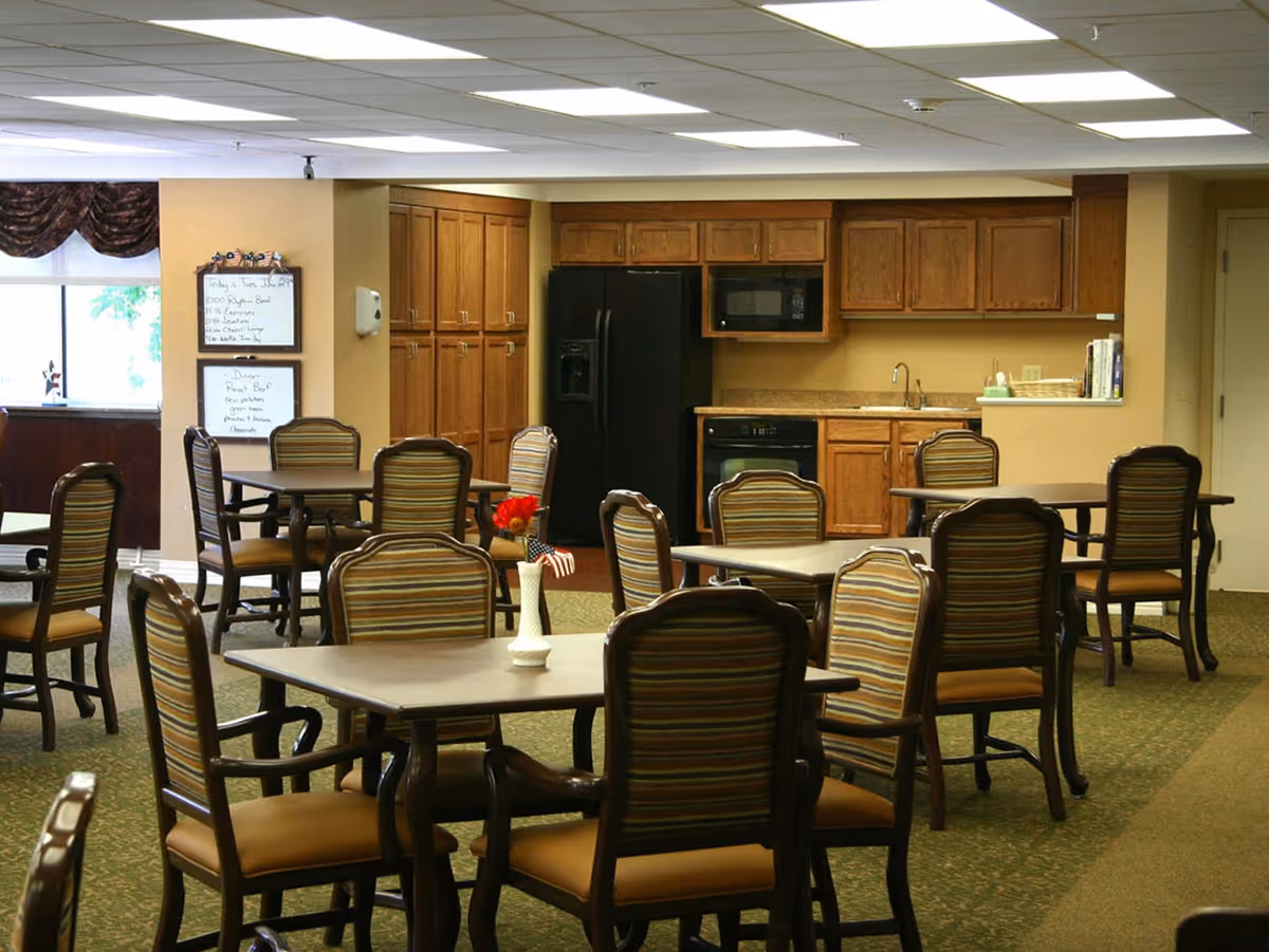 Dining area with multiple tables and chairs featuring striped upholstery. In the background, there is a kitchen area with wooden cabinets, a black refrigerator, a microwave, a dishwasher, and a sink. A window with dark valance curtains and two whiteboards with handwritten notes are visible on the left wall.