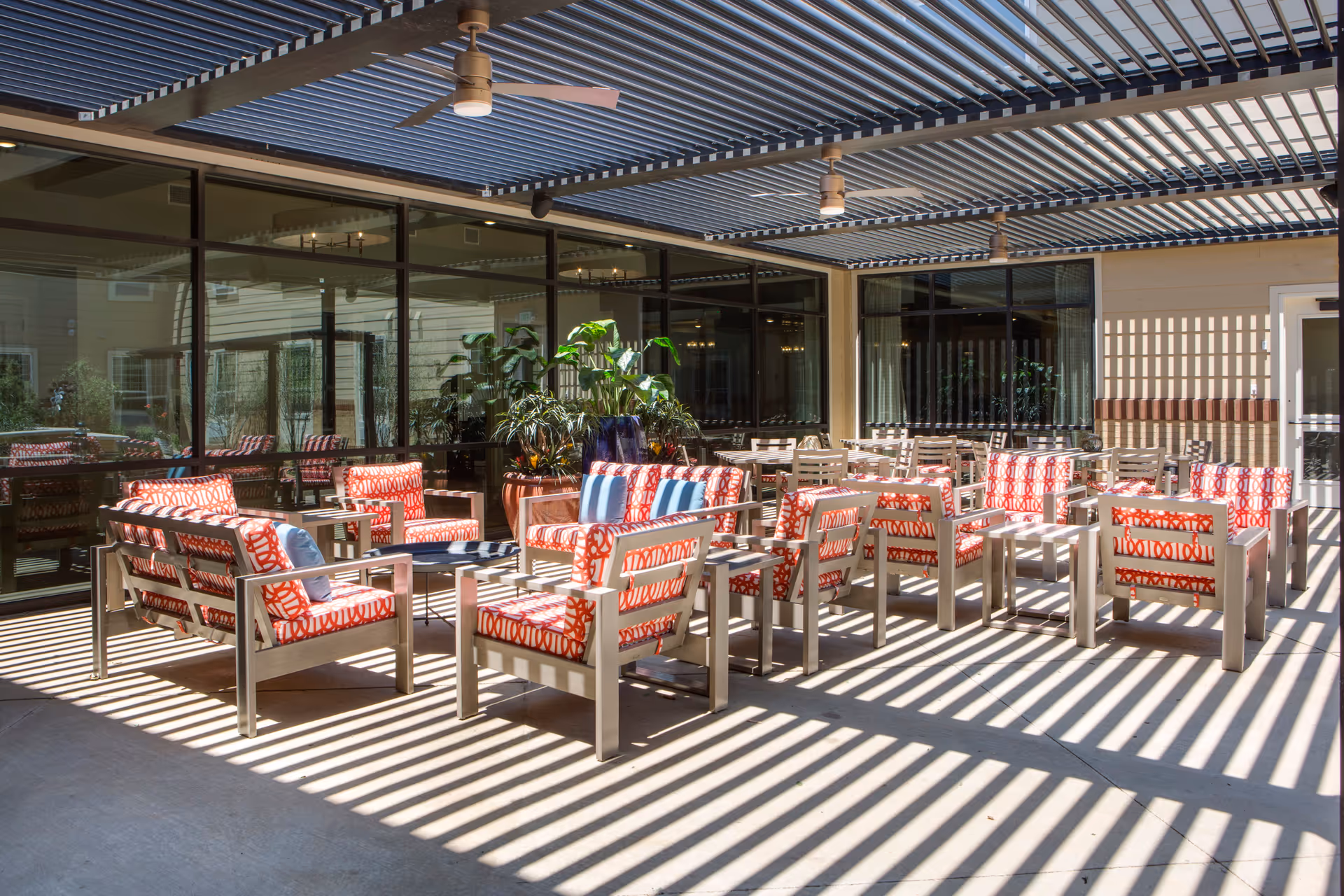 Outdoor patio area with modern seating featuring red and white patterned cushions, several tables and chairs, potted plants, and a slatted pergola casting striped shadows on the concrete floor.