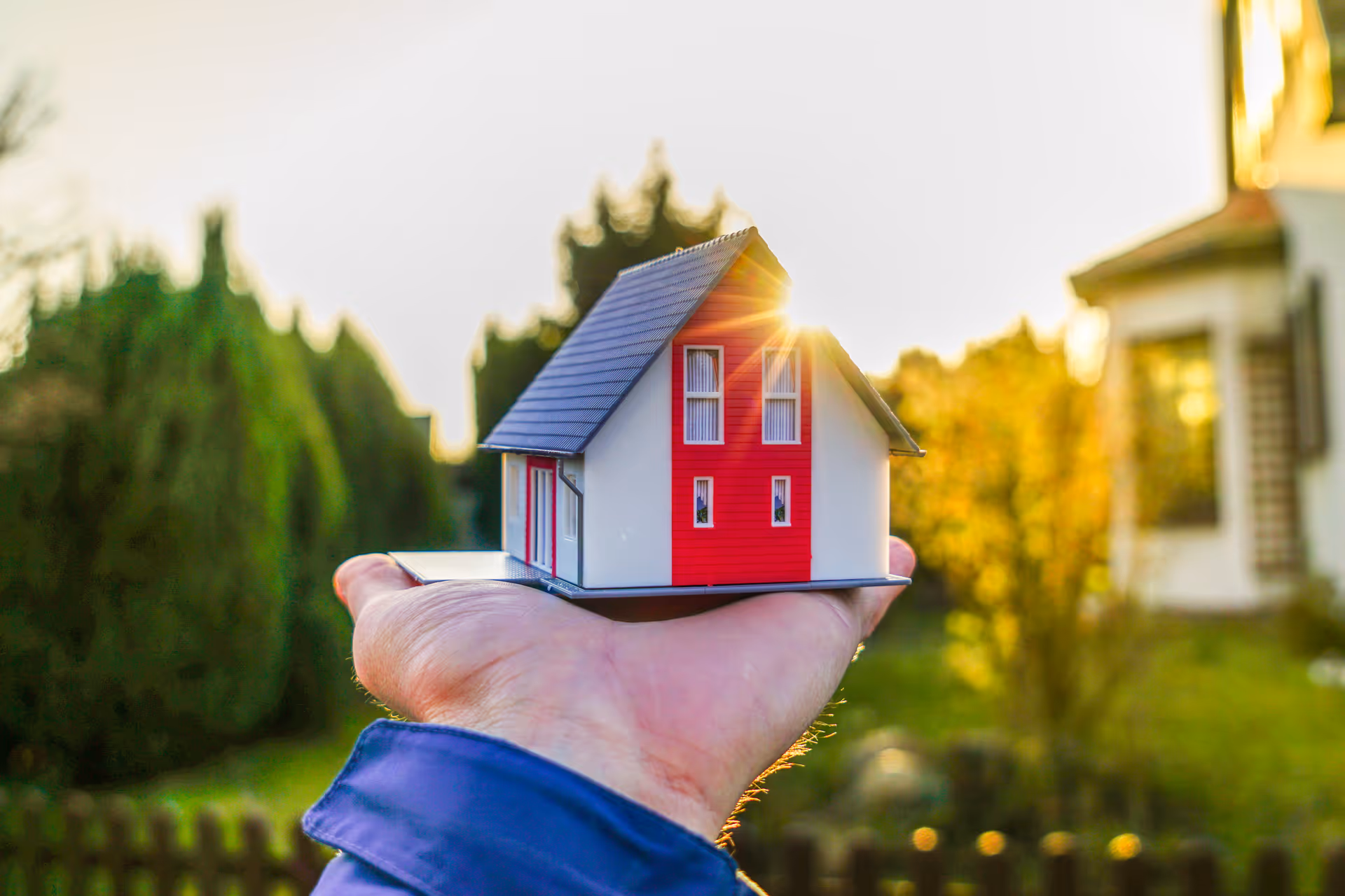 A hand holding a small model house with a red facade outdoors at sunset.