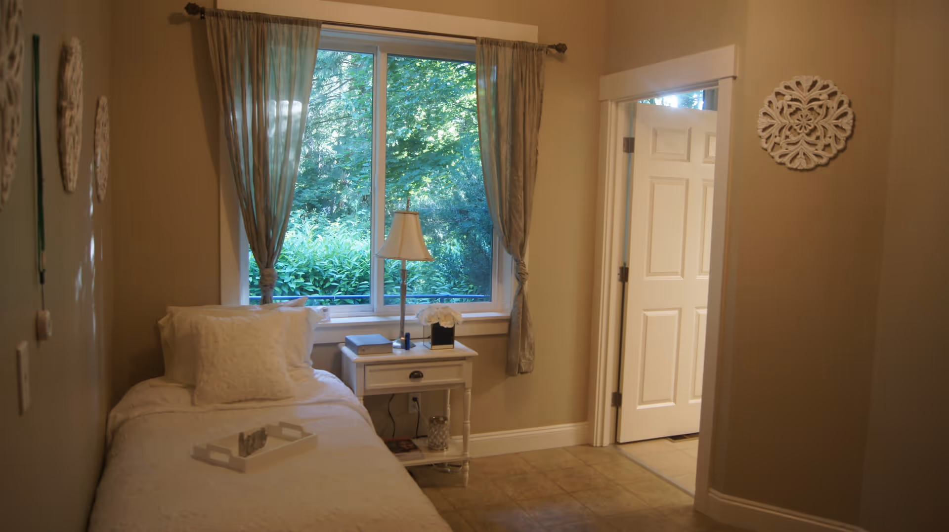 Single-occupancy bedroom with a twin bed, white bedside table and lamp under a large window looking out to greenery.