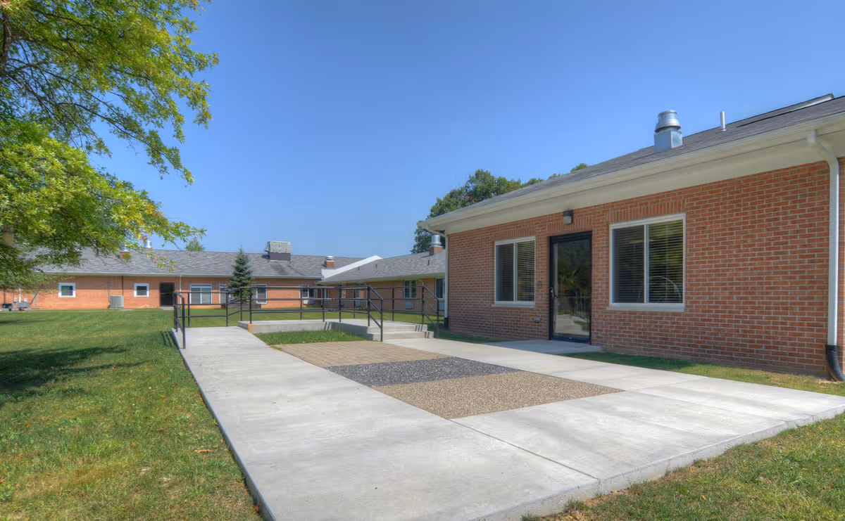 Exterior view of a single-story brick building with a concrete walkway and ramp leading to a glass door entrance. The building is surrounded by green grass and trees under a clear blue sky.