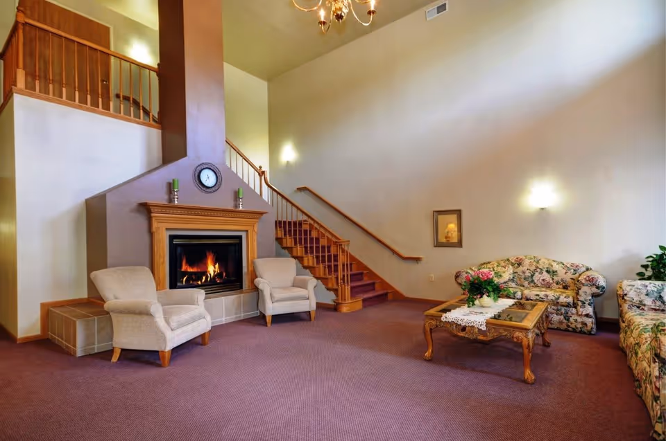 A cozy living room area with a lit fireplace, two beige armchairs, a floral-patterned sofa and loveseat, a wooden coffee table with a lace runner and flower arrangement, and a staircase with wooden railing leading to an upper floor.