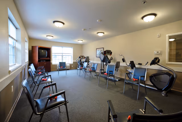 A fitness room in a senior living facility with several exercise machines including stationary bikes and treadmills. There are black chairs arranged in a circle, each with a pair of red hand weights on the seat. The room has beige walls, carpeted floor, and ceiling lights. A wooden cabinet with a television is positioned near the windows letting in natural light.