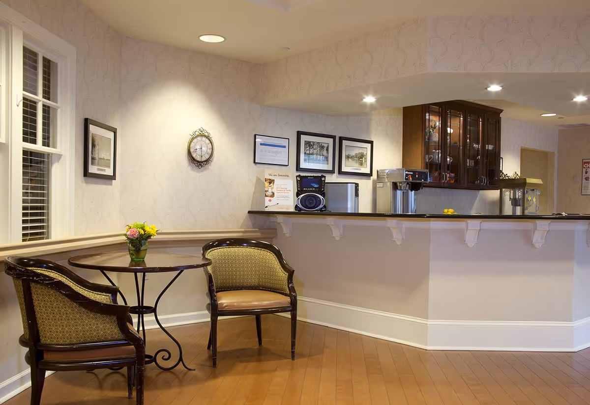 A cozy seating area in a senior living facility with two upholstered chairs around a small round table holding a vase of flowers. Behind the seating area is a counter with a coffee machine and a popcorn maker, with cabinets and framed pictures on the wall.