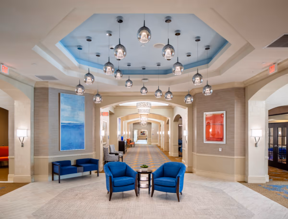 A spacious and well-lit hallway in a senior living facility with a high ceiling featuring multiple hanging pendant lights. The hallway has a blue and beige color scheme with blue armchairs and a small round table in the foreground. There are artworks on the walls and several seating areas along the corridor.