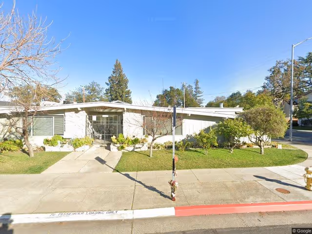 Single-story building with a flat roof and large windows, surrounded by a small lawn with bushes and trees, located on a street corner under a clear blue sky.