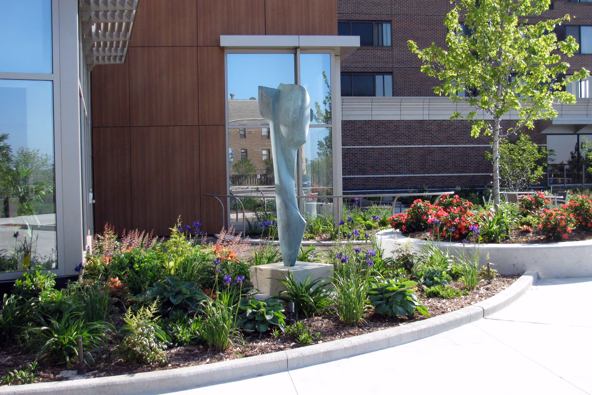 Outdoor garden area at Saint John's On The Lake featuring a modern abstract sculpture surrounded by various green plants and colorful flowers, with a tree and a building in the background.