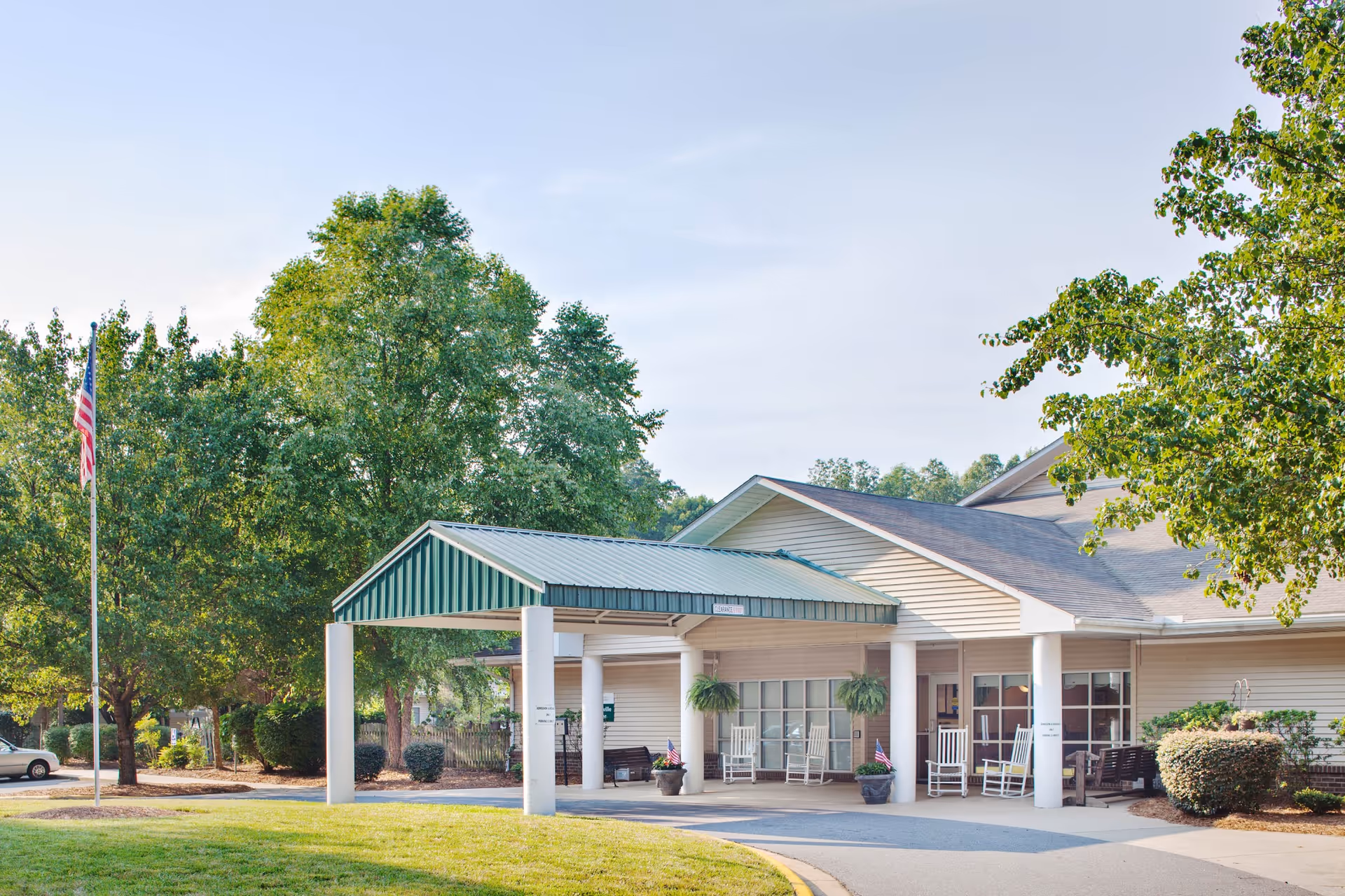 Exterior view of Mooresville Center showing the entrance with a covered drop-off area supported by white columns. There are rocking chairs and potted plants near the entrance. The building is surrounded by green trees and bushes, with an American flag on a flagpole to the left.