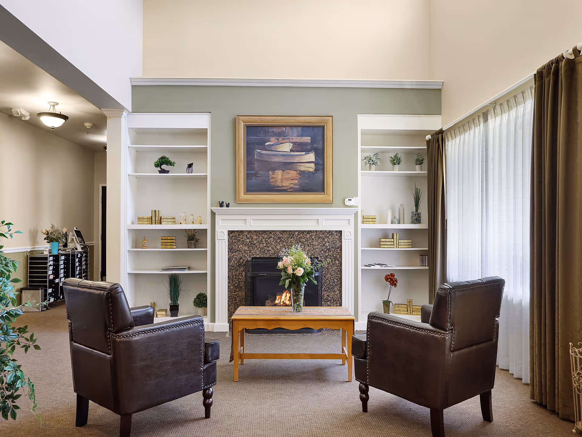 A cozy living room area with two dark brown leather armchairs facing a wooden coffee table with a vase of flowers. Behind the table is a fireplace with a granite surround and a painting of boats above it. White built-in shelves flank the fireplace, decorated with books and small plants. Large windows with sheer white curtains and brown drapes allow natural light into the room.
