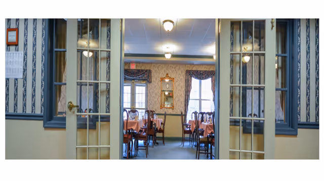 Interior view through glass-paned double doors into a formal dining room with tables set with pink tablecloths, wooden chairs, wallpaper, and draped windows.