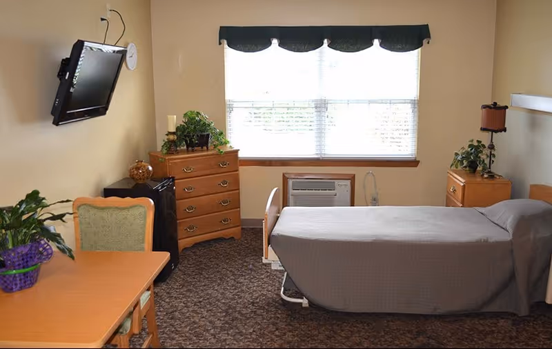 A small bedroom in a senior living facility with a single bed covered in a gray blanket, a wooden dresser, a nightstand with a lamp and plant, a wall-mounted TV, a table with a chair, and a window with blinds and a dark valance.