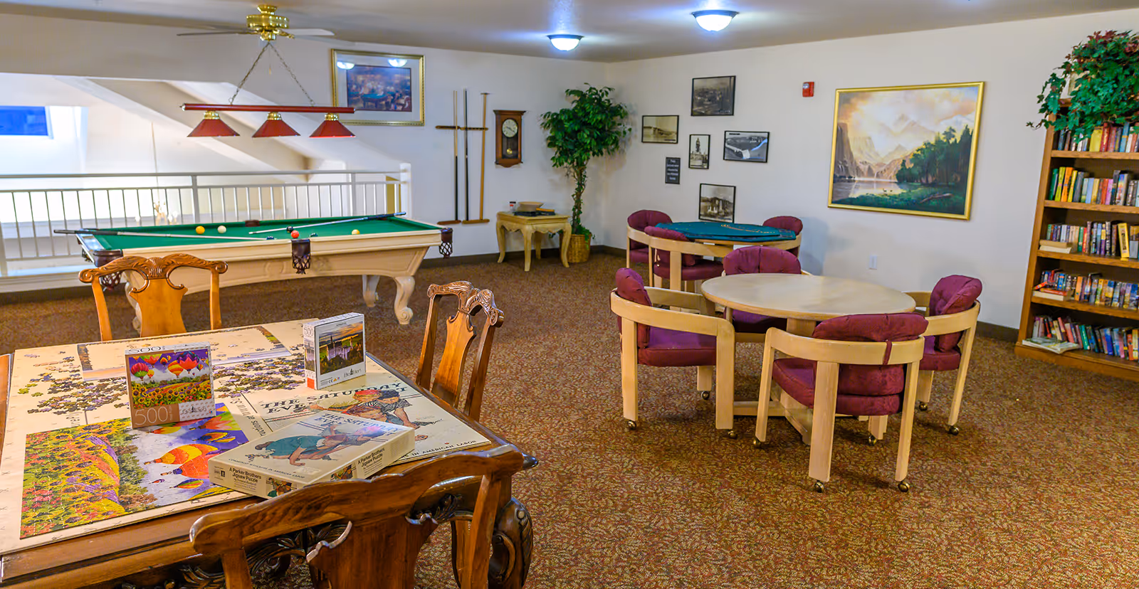 A recreational room with a pool table, a card table with four purple cushioned chairs, a wooden table with chairs and several jigsaw puzzles on it, a bookshelf filled with books, and framed pictures and a clock on the walls. The room has carpeted flooring and ceiling lights.