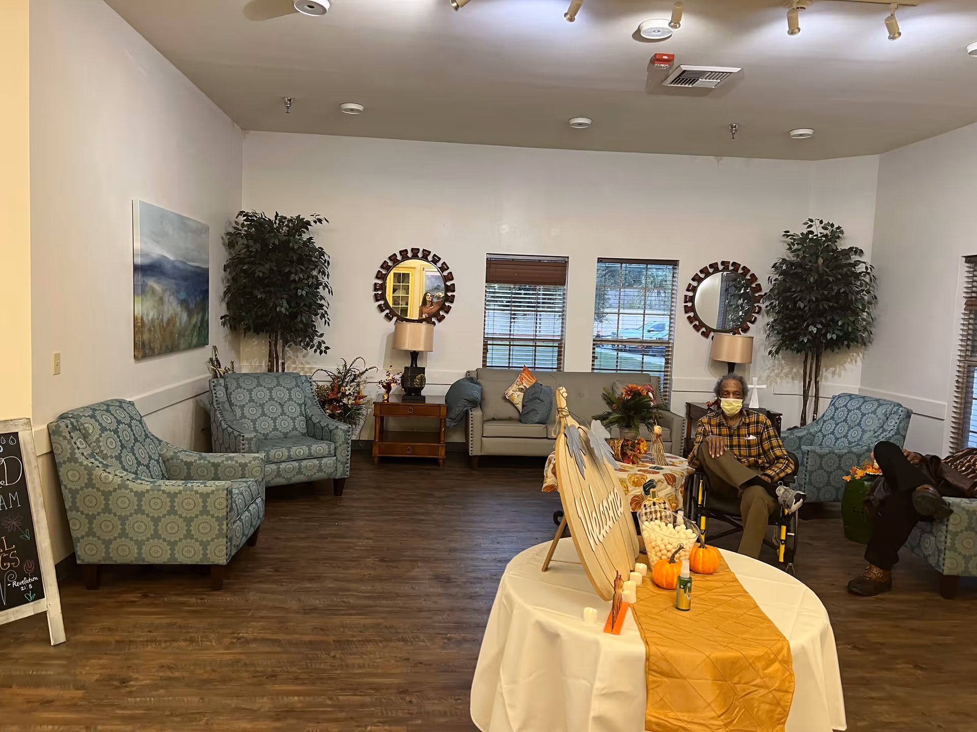 A senior living community lounge with upholstered chairs and sofas around a decorated table, with residents seated (one in a wheelchair).