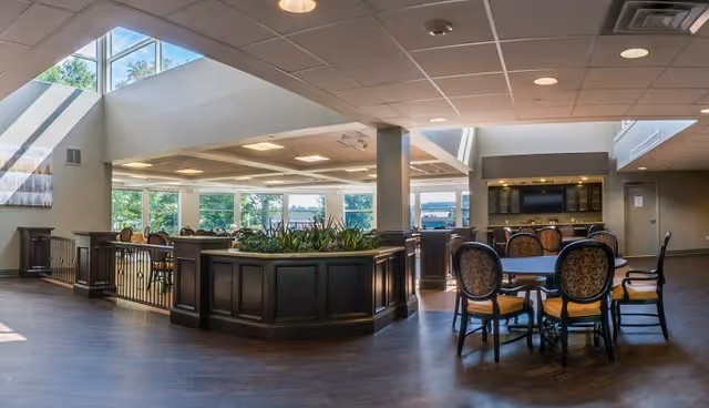 A spacious and well-lit interior common area with large windows allowing natural light to fill the room. There are several round tables with cushioned chairs arranged around them, and a central planter with greenery. The ceiling has recessed lighting and a modern design, and the flooring is dark wood.