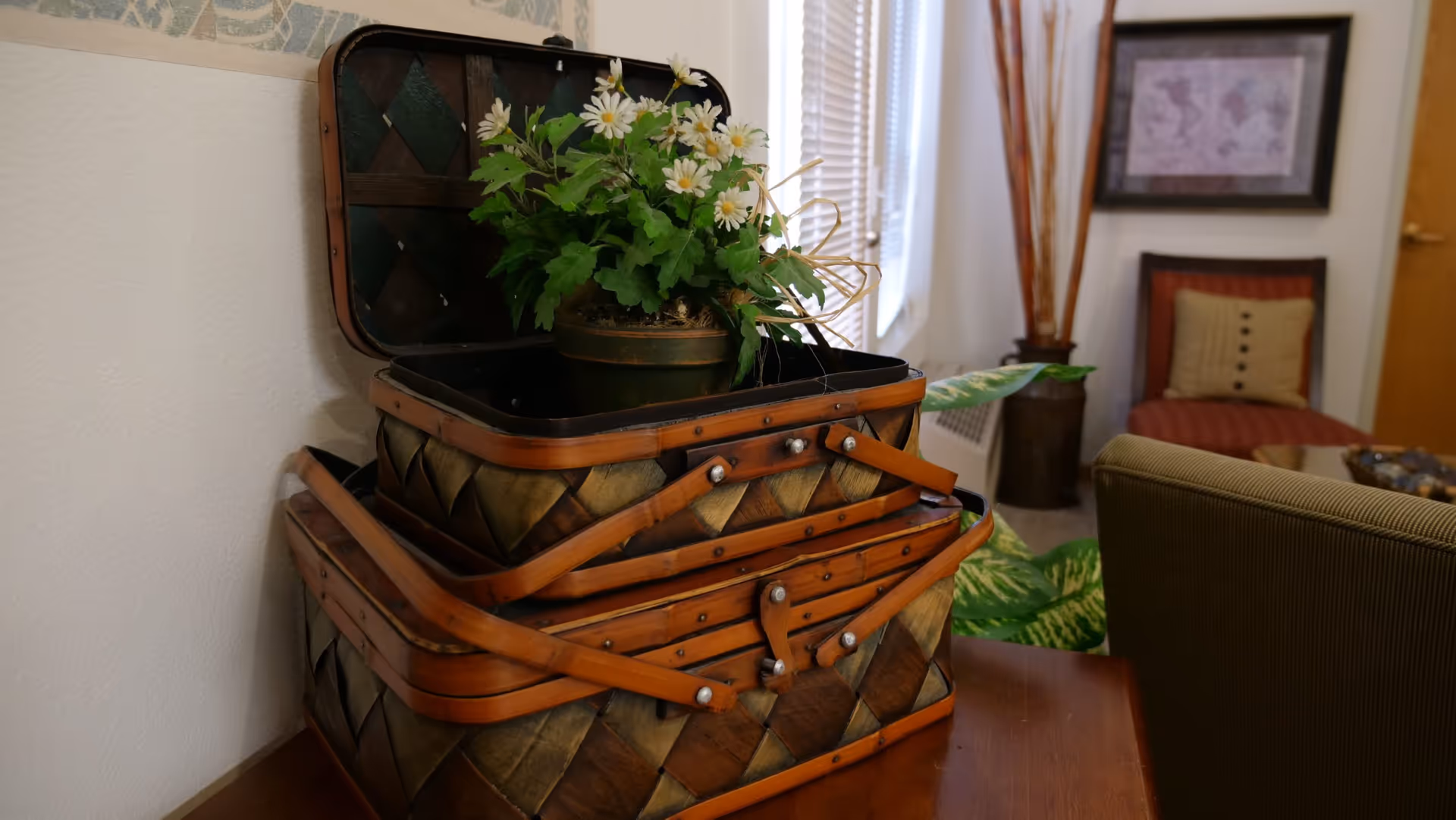A decorative arrangement featuring a potted plant with white flowers placed inside an open woven basket on a wooden table. In the background, there is a red cushioned chair with a beige pillow, a framed map on the wall, and a tall vase with dried branches near a window with blinds.