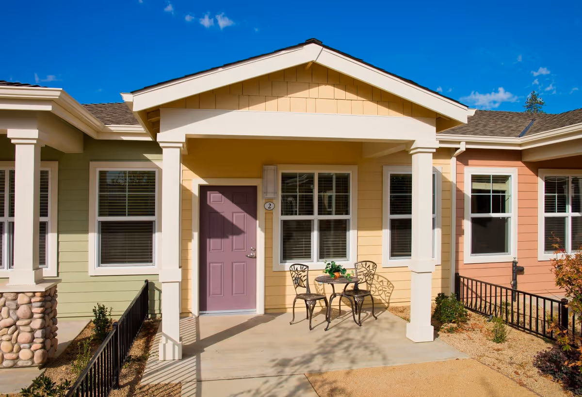 Exterior view of a single-story residential unit with a covered porch featuring a small round metal table and two matching chairs. The building has light green, yellow, and peach siding with white trim around windows and doors. The door is painted purple and marked with the number 2. The porch area is surrounded by small plants and a black metal fence under a clear blue sky.