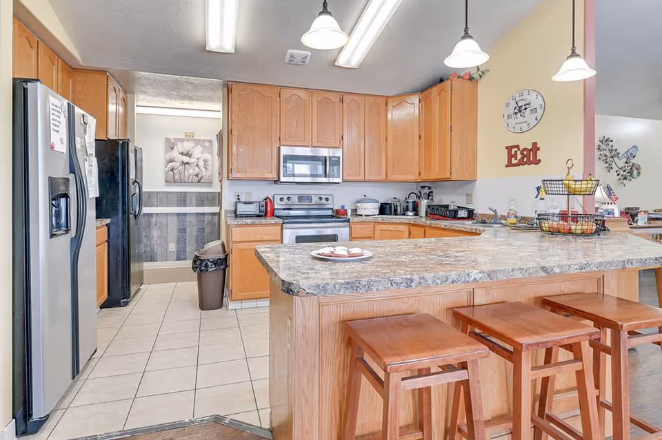 A bright kitchen with wooden cabinets and a large island countertop with three wooden stools. The kitchen features a stainless steel refrigerator, microwave, and stove. There are three pendant lights hanging above the island. On the wall, there is a clock and a decorative sign that says 'Eat'. The floor is tiled, and there is a trash bin near the entrance to another room with a floral wall decoration.