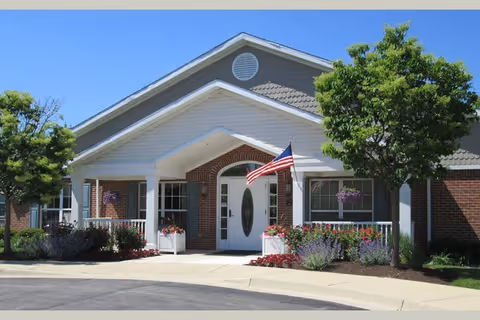 Front exterior view of a single-story brick building with a white entrance porch, an American flag mounted near the door, and landscaping including trees and flower beds under a clear blue sky.