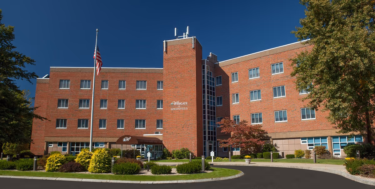 Exterior view of a four-story brick building with multiple windows, an American flag on a flagpole in front, and landscaped greenery including bushes and trees under a clear blue sky. The building has a sign that reads 'Wingate Springfield' near the entrance.