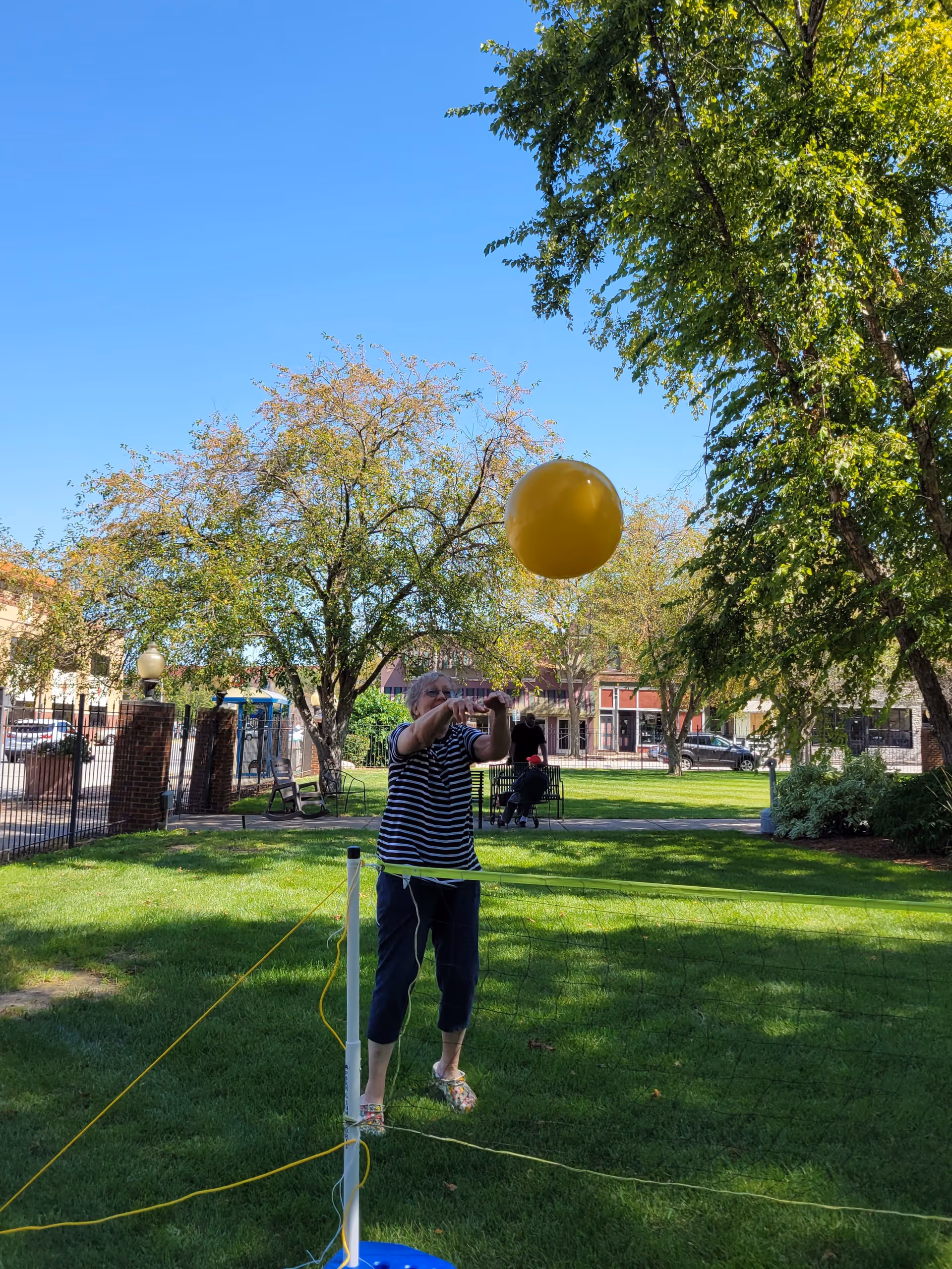 An elderly woman playing with a yellow balloon over a small net in a grassy outdoor area with trees and buildings in the background under a clear blue sky.