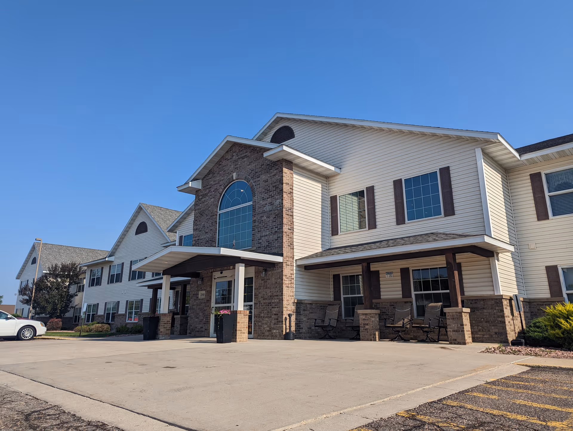 Exterior view of a two-story senior living facility building with beige siding and brick accents under a clear blue sky. The entrance has a covered porch with seating and potted flowers, and there is a parking area in front with a white car parked to the left.