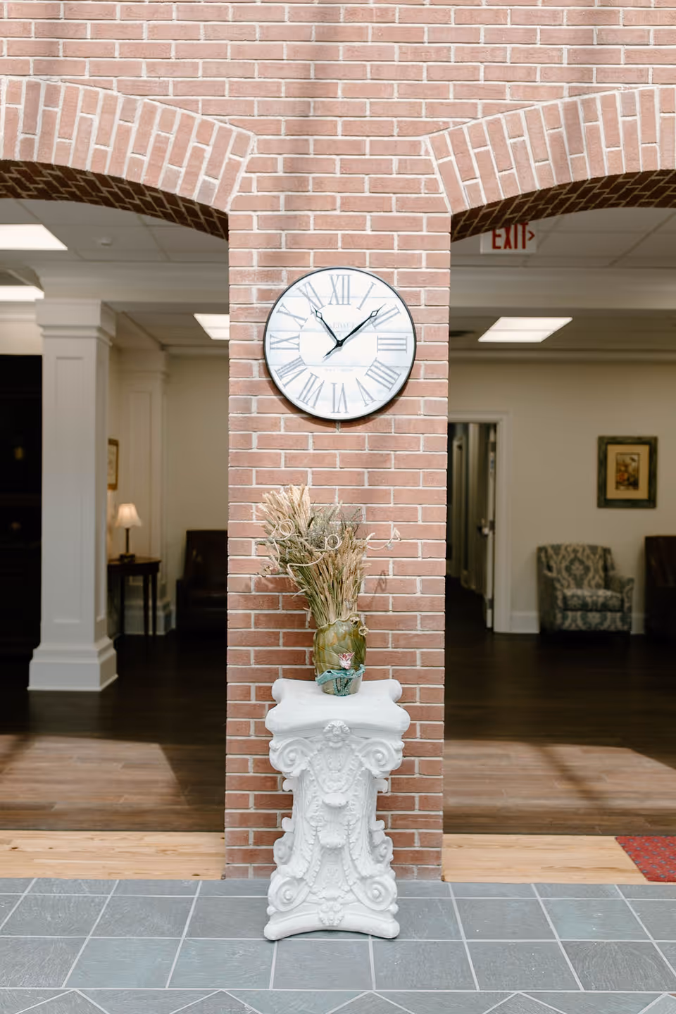 Interior view of a senior living facility showing a brick column with a round clock displaying the time 11:10. Below the clock is a decorative white pedestal with a vase containing dried plants. The background includes two archways leading to a room with chairs, a lamp, framed artwork, and an exit sign on the ceiling.
