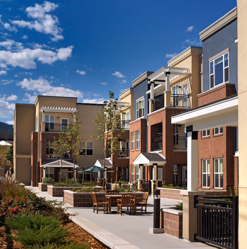 Outdoor courtyard area of a senior living facility with multi-story buildings featuring balconies, patio tables with chairs, landscaped garden beds, and a clear blue sky with scattered clouds.