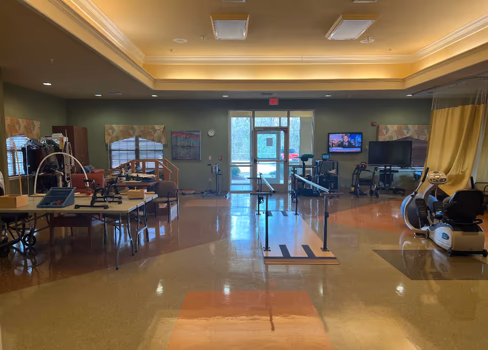 A spacious rehabilitation or physical therapy room with parallel bars in the center for walking exercises, exercise equipment including a recumbent bike on the right, and tables with various therapy tools on the left. The room has a polished floor, green walls, windows with patterned valances, and a TV mounted on the wall. A glass door at the back leads outside.