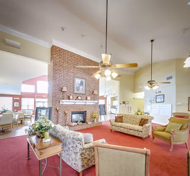 A spacious living room area with a red carpet, multiple upholstered sofas and chairs arranged around a brick fireplace. The room has high ceilings with ceiling fans and light fixtures. There is a wooden side table with a floral arrangement, and large windows in the background letting in natural light. The adjacent dining area is visible through an open doorway.