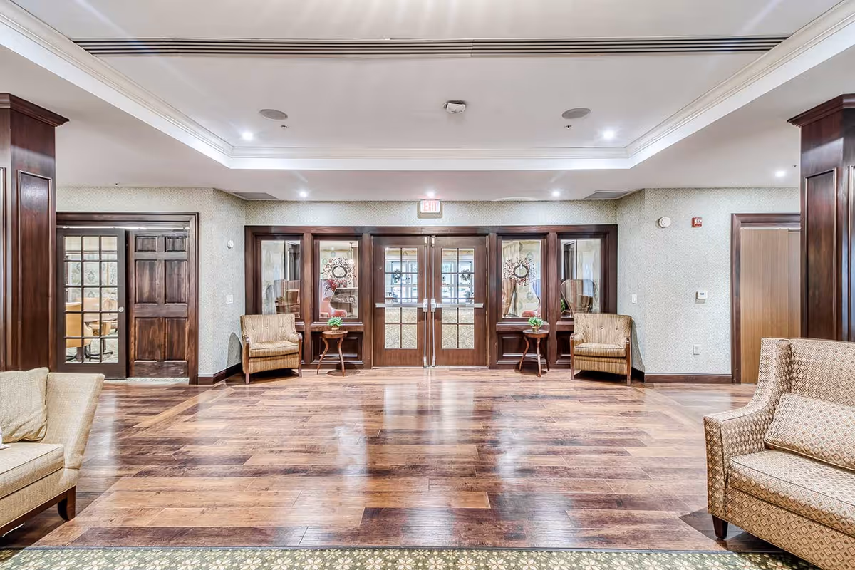 A spacious, well-lit interior common area with polished wooden floors and beige patterned walls. The room features several cushioned armchairs and small wooden tables with plants. At the far end, there are double glass doors with wooden frames, flanked by additional glass panels and chairs. The ceiling has recessed lighting and a decorative tray design.