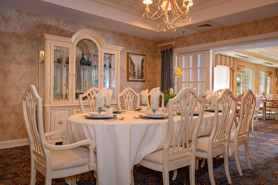 A formal dining room with a large oval table covered with a white tablecloth, set with plates, glasses, and napkins. The room features elegant wooden chairs with cushioned seats, a glass-fronted cabinet displaying decorative glassware, a chandelier overhead, and a painting on the wall. In the background, there is another dining area visible through a wide doorway.
