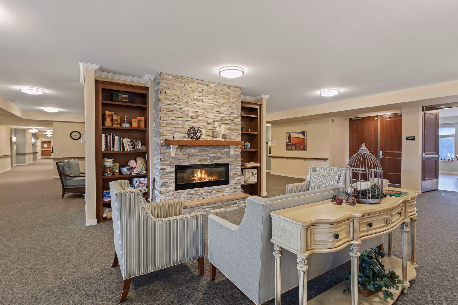 A cozy seating area in a senior living facility featuring a stone fireplace with a wooden mantel, two striped armchairs, a light-colored sofa, and a decorative table with a birdcage and plants. Bookshelves flank the fireplace, and a hallway with doors and wall art is visible in the background.