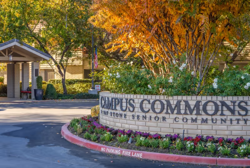 Entrance to Campus Commons senior living community with a brick sign surrounded by flowers and trees with autumn foliage. A covered drop-off area is visible in the background.