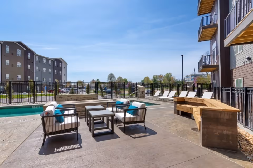 Outdoor patio area with cushioned chairs and small tables arranged near a swimming pool, surrounded by a black metal fence. Several lounge chairs are lined up along the poolside, and there is a wooden bench on the right side. Apartment buildings and trees are visible in the background under a clear blue sky.