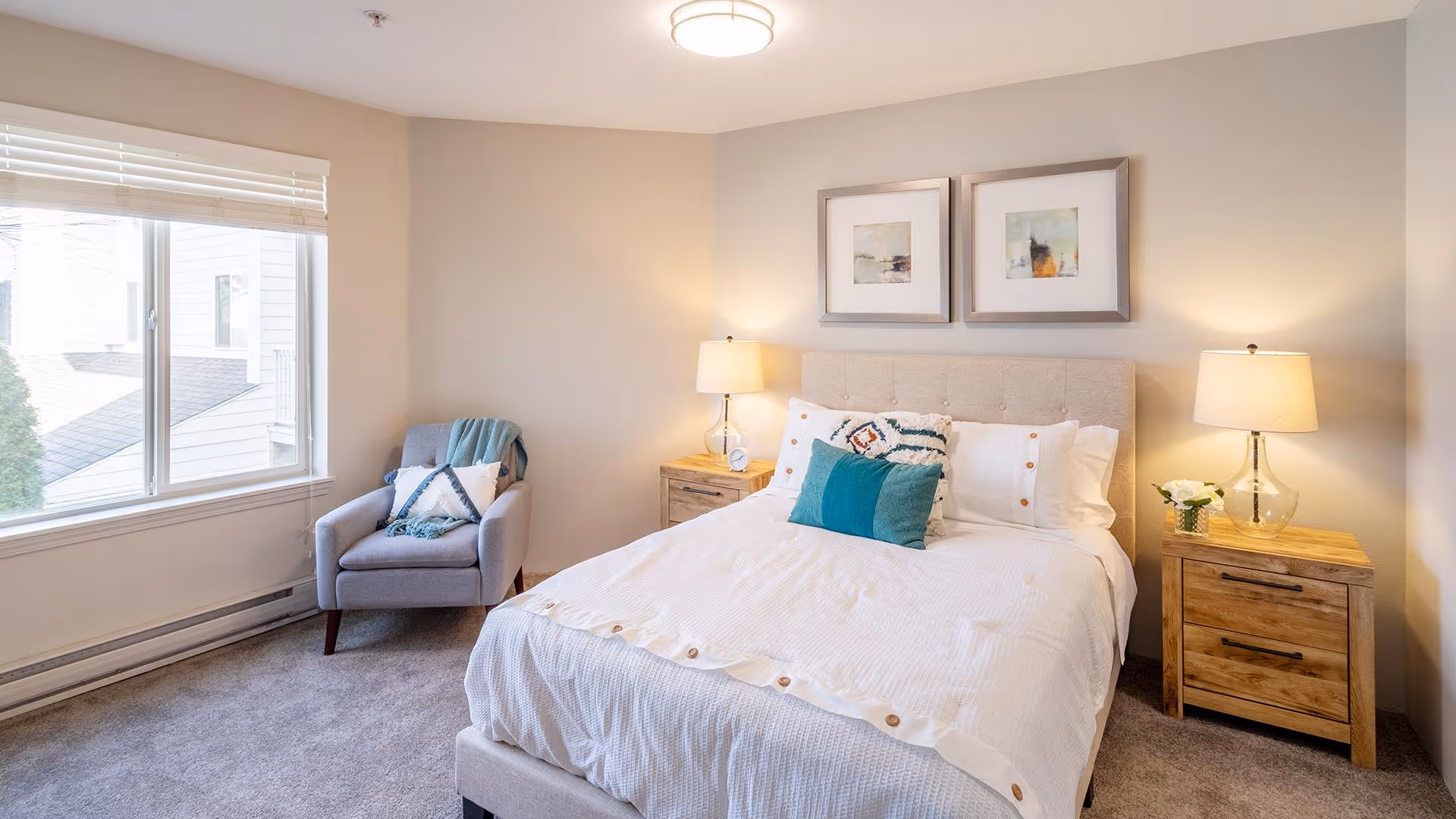 A bright and cozy bedroom with a large bed featuring white bedding and decorative pillows. On either side of the bed are wooden nightstands with lamps and a small flower arrangement on the right nightstand. A comfortable gray armchair with a throw blanket and pillow is placed near a large window with white blinds. Two framed abstract artworks hang above the bed.