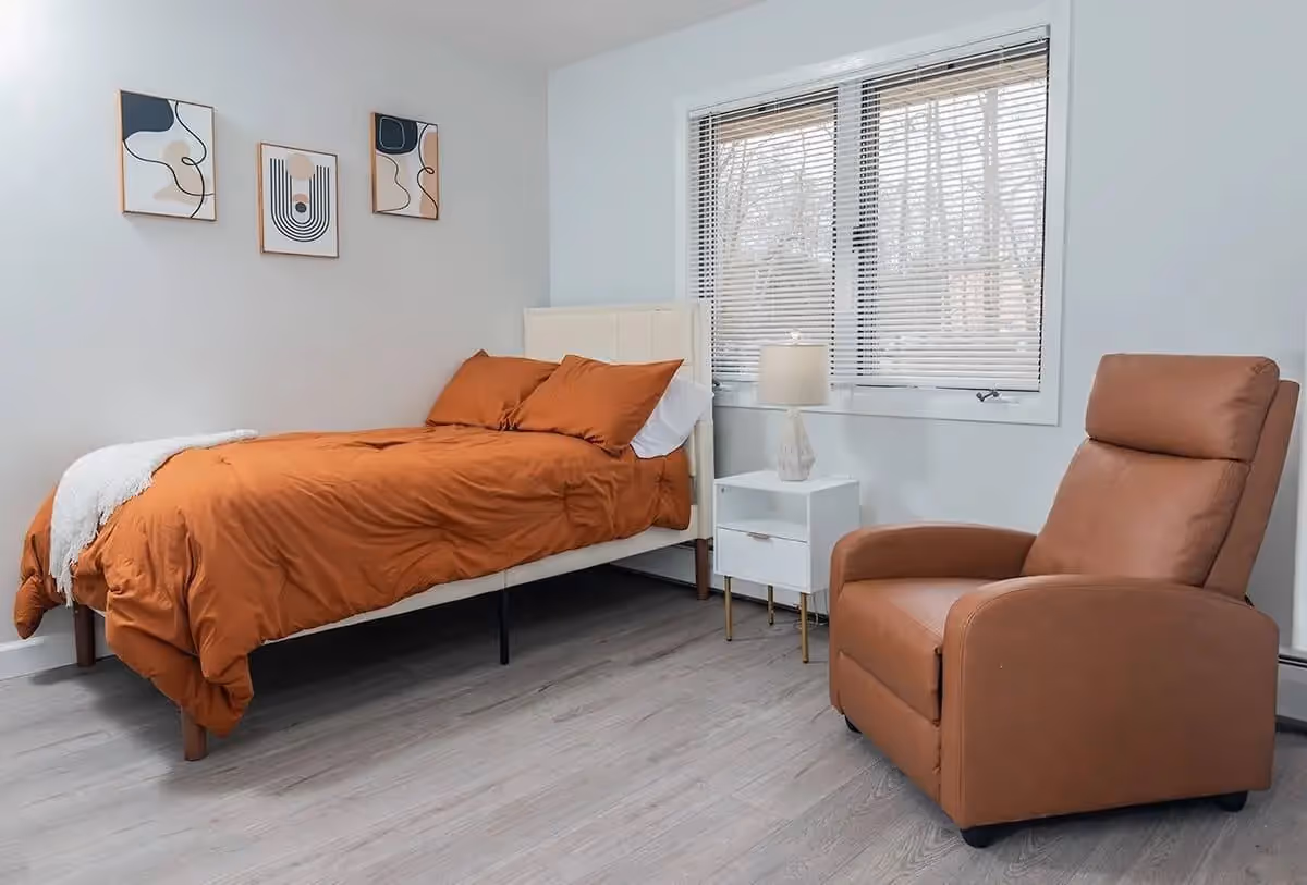 A bedroom with a single bed dressed in orange bedding and a white throw blanket. Next to the bed is a white nightstand with a lamp. A large window with blinds is behind the nightstand. There is a brown recliner chair to the right of the nightstand. Three abstract art pieces hang on the wall above the bed. The floor is light-colored wood.