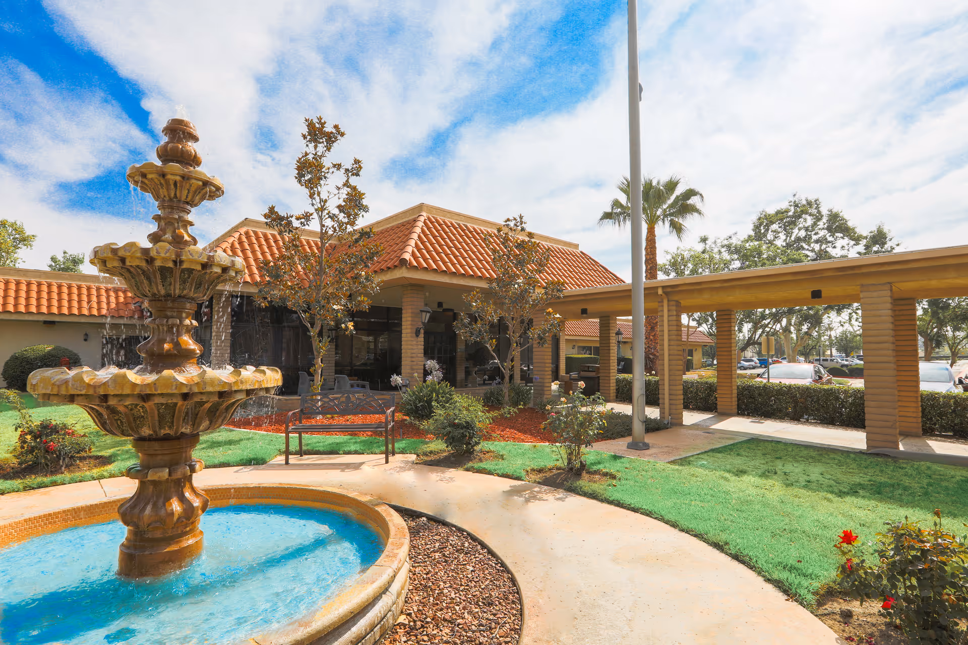 Outdoor view of Corona Post Acute facility featuring a multi-tiered water fountain in the foreground, a curved walkway, green lawn, small trees, and a building with a red tiled roof under a partly cloudy blue sky.