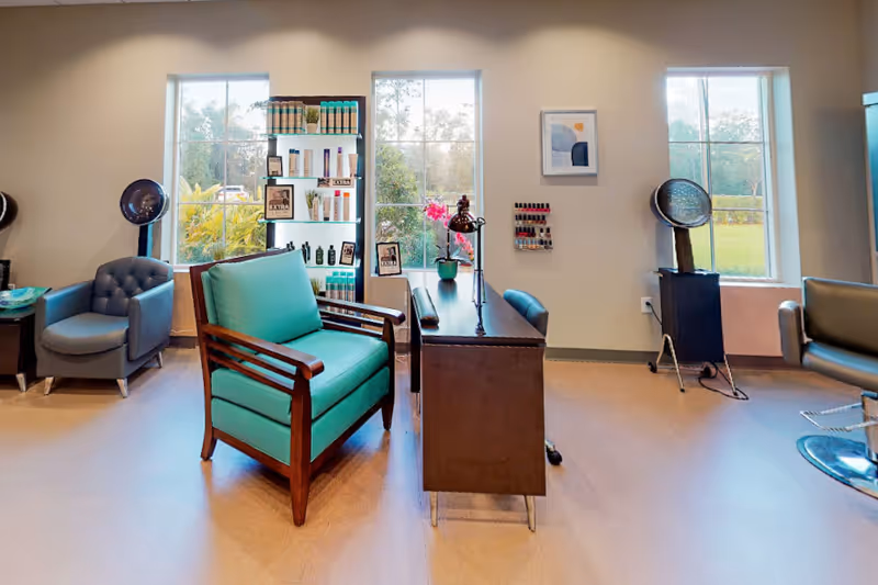 Bright salon-style interior with a teal armchair, manicure table, hair-dryer stations and product shelving in front of three windows.
