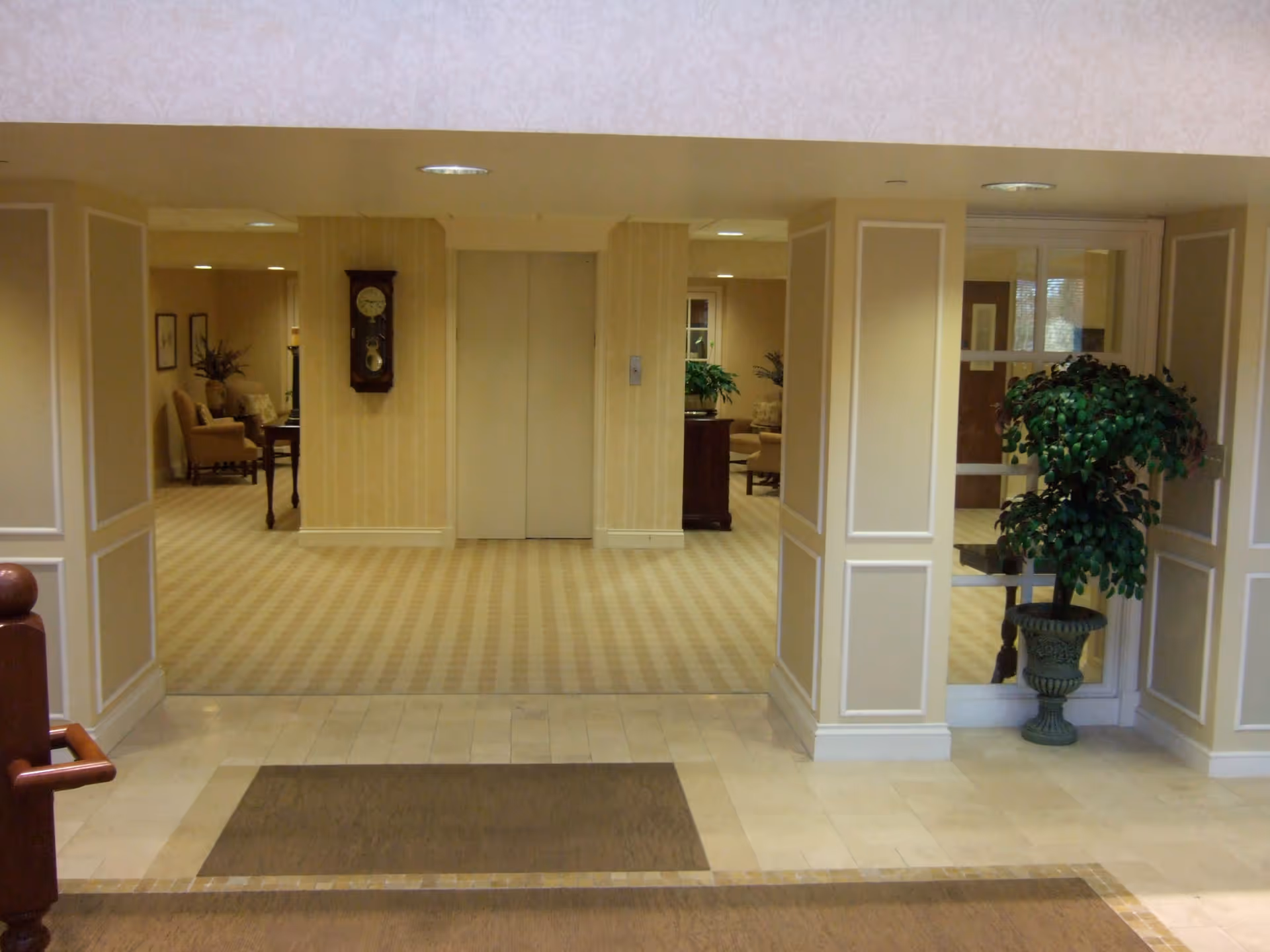 Interior view of a senior living facility hallway with beige patterned carpet and wallpaper. There are two elevator doors in the center with a wooden wall clock mounted between them. On either side of the hallway, there are seating areas with armchairs, tables, and plants. A large potted plant is placed near a glass door on the right side. The floor near the entrance has light-colored tiles with a brown mat.
