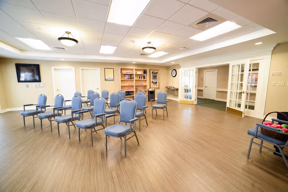 A spacious room with multiple blue cushioned chairs arranged in rows on a wooden floor. The room has beige walls, a white ceiling with recessed lighting and ceiling fixtures, a water fountain, a clock on the wall, and a bookshelf with various items. There are double glass doors on the right side and two closed white doors on the back wall. A basket with colorful balls is placed on a chair in the right foreground.