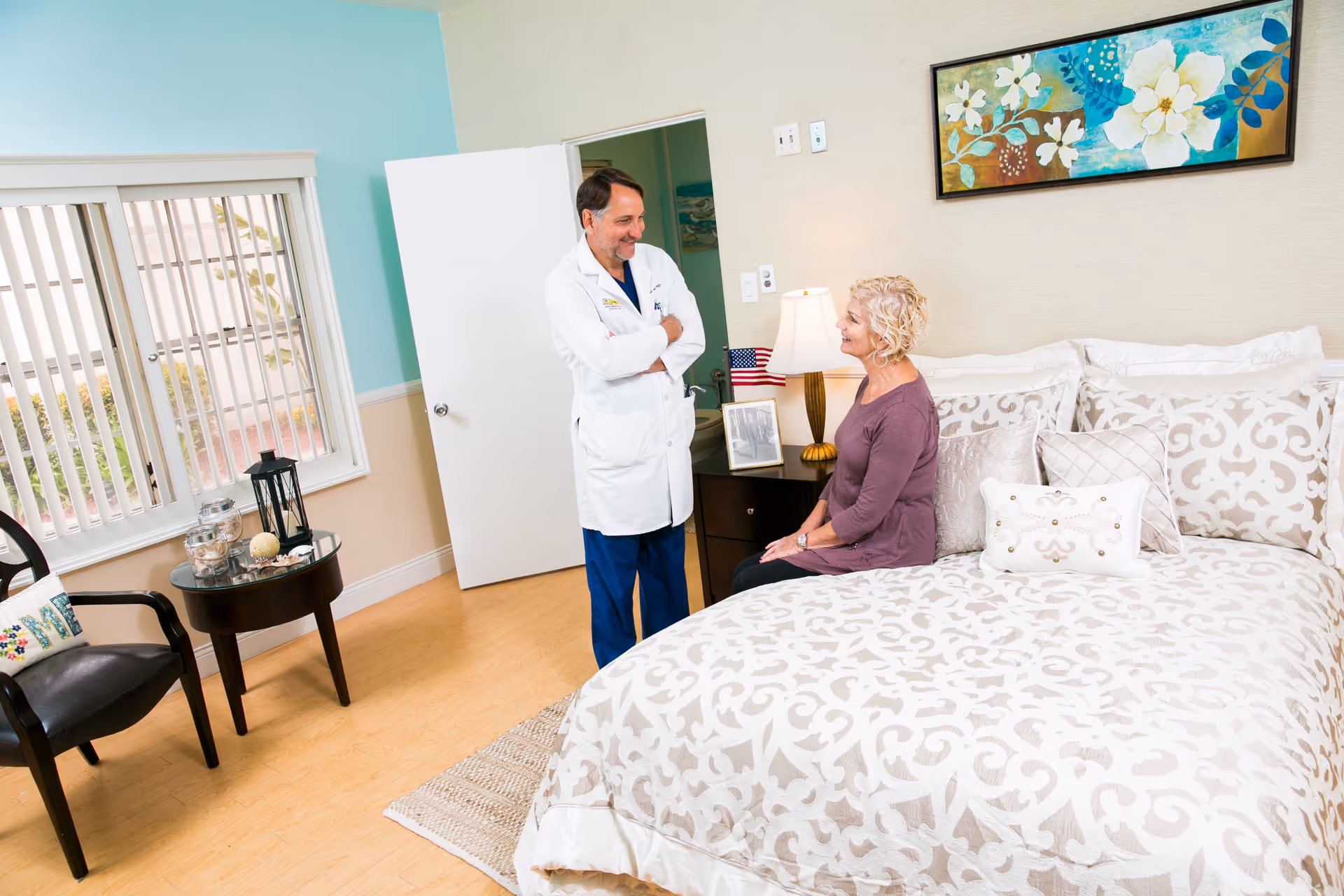 A doctor in a white coat is standing and talking with a seated elderly woman in a well-lit bedroom. The room features a large bed with patterned bedding, a bedside table with a lamp and small American flag, a chair, and a small round table with decorative items. A colorful floral painting hangs on the wall above the bed.