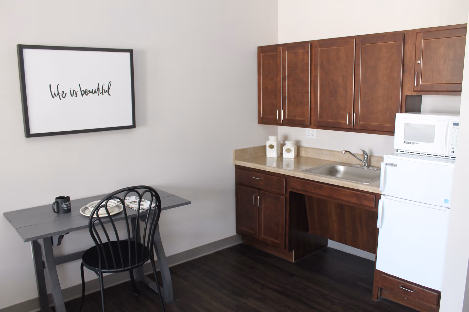 A small kitchen area with dark wooden cabinets, a countertop with a sink, a white microwave on top of a white mini refrigerator, and a small dining table with a black chair. On the wall above the table is a framed sign that reads 'life is beautiful'.