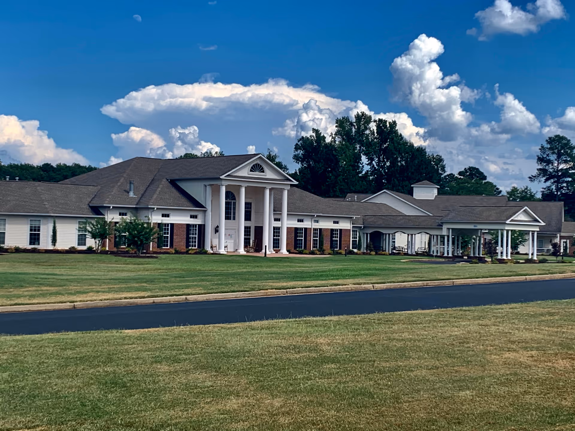 Exterior view of a single-story senior living facility building with white columns at the entrance, surrounded by a well-maintained lawn and trees under a partly cloudy blue sky.