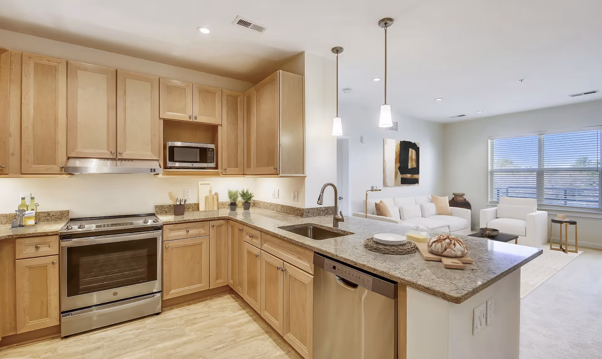 Open modern kitchen with light wood cabinets, a granite island and stainless steel appliances opening to a bright living room.