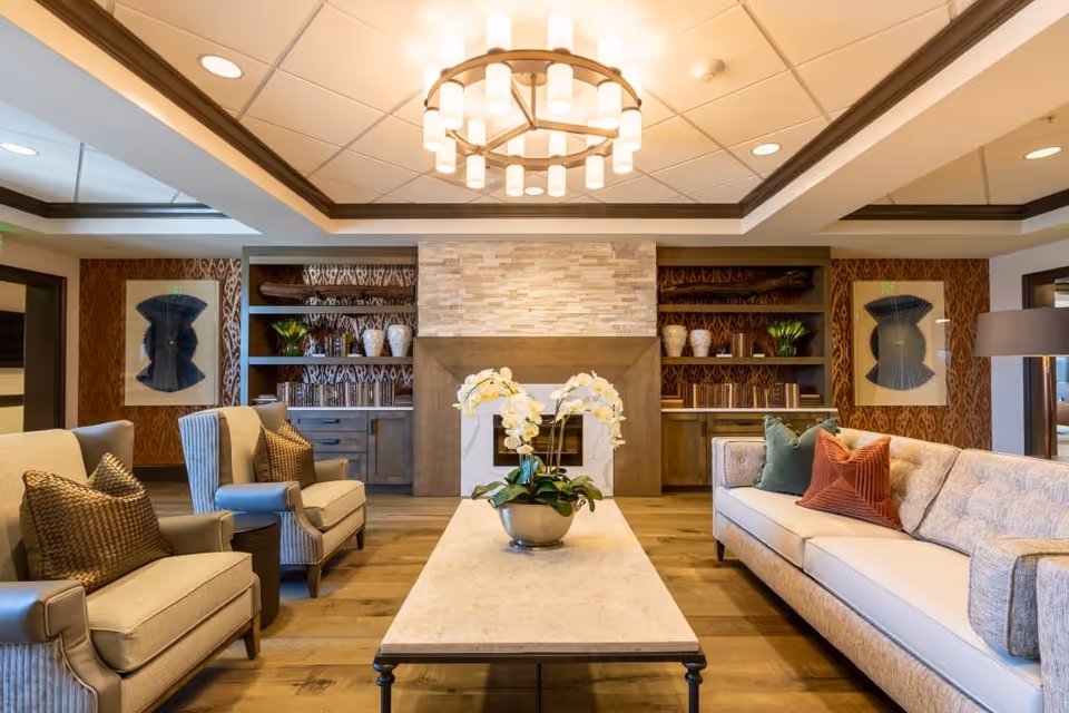 A cozy living room with a beige sofa adorned with green and rust-colored pillows on the right, and two beige armchairs with brown cushions on the left. A rectangular coffee table with a potted white orchid sits in the center. The background features a stone fireplace flanked by built-in wooden shelves decorated with vases, books, and plants. Two framed abstract artworks hang on the patterned wallpapered walls. A modern circular chandelier hangs from the ceiling.
