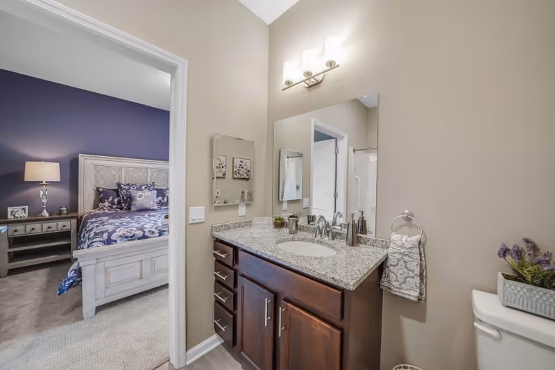 Bathroom vanity with granite countertop, sink, mirror and light fixture, with a bedroom visible through the open doorway.