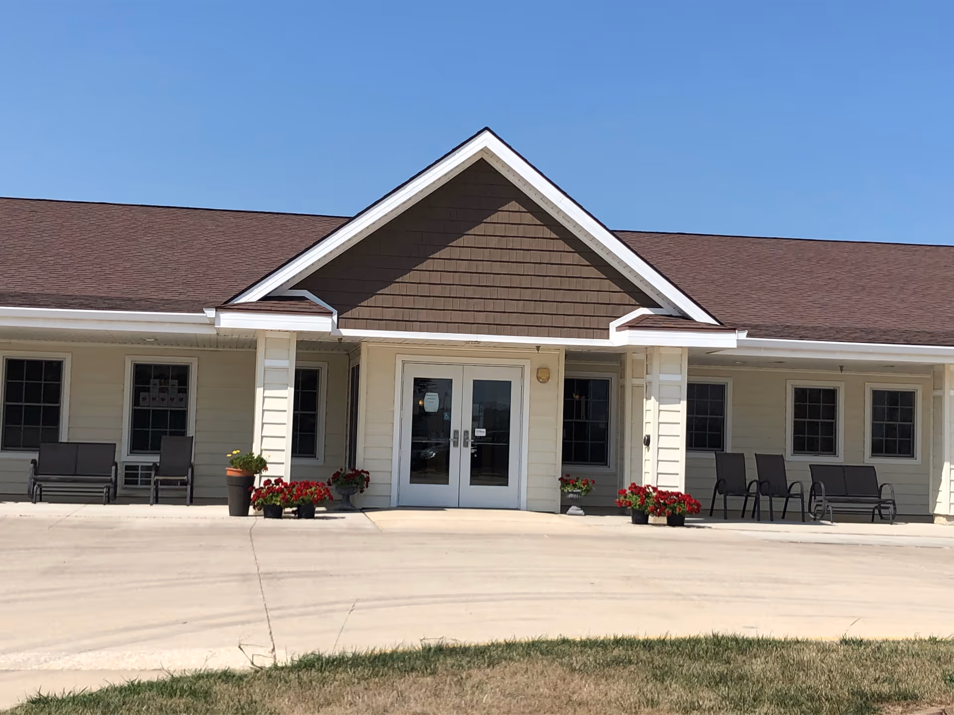 Front exterior view of The Gardens Assisted Living facility with a peaked roof, double glass entrance doors, several windows, outdoor seating benches, and flower pots with red flowers on a sunny day.