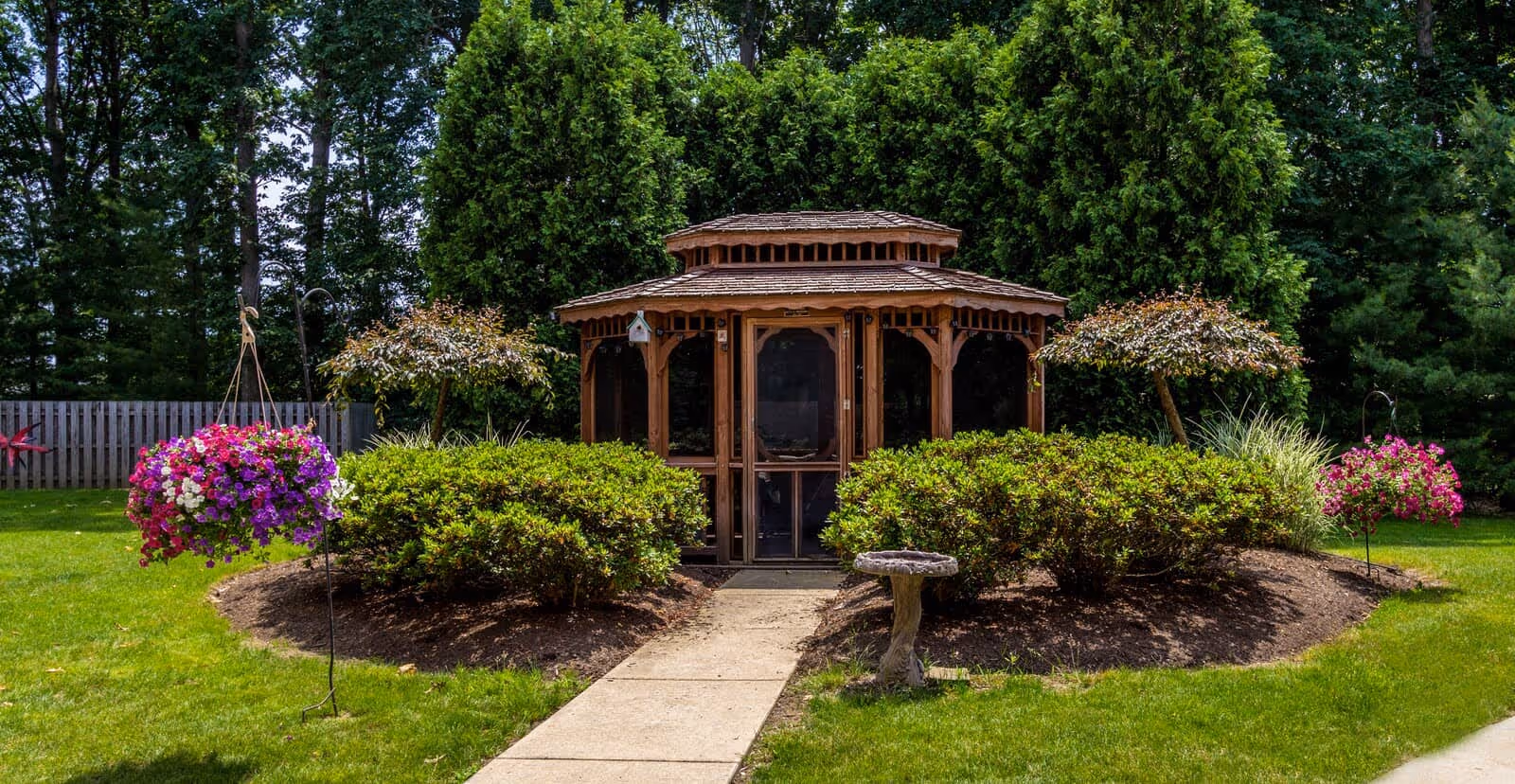 A wooden gazebo centered in a landscaped lawn surrounded by shrubs, hanging flower baskets, and trees.
